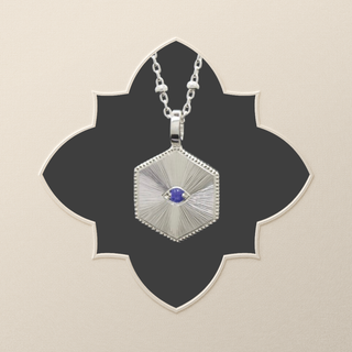 A silver hexagonal pendant necklace with a central blue gemstone, displayed on a black four-petal cutout over a light beige background.