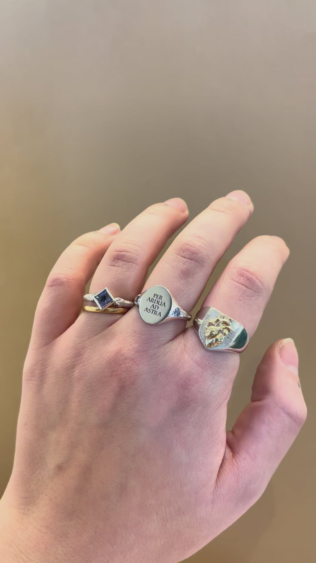 Hand wearing three rings, a silver shield signet with an anatomical human heart in gold, a silver latin motto engraved oval signet ring with birthstone set band, a princess cut gemstone solitaire engagement ring with a blue sapphire with snake head band - emily proudfoot