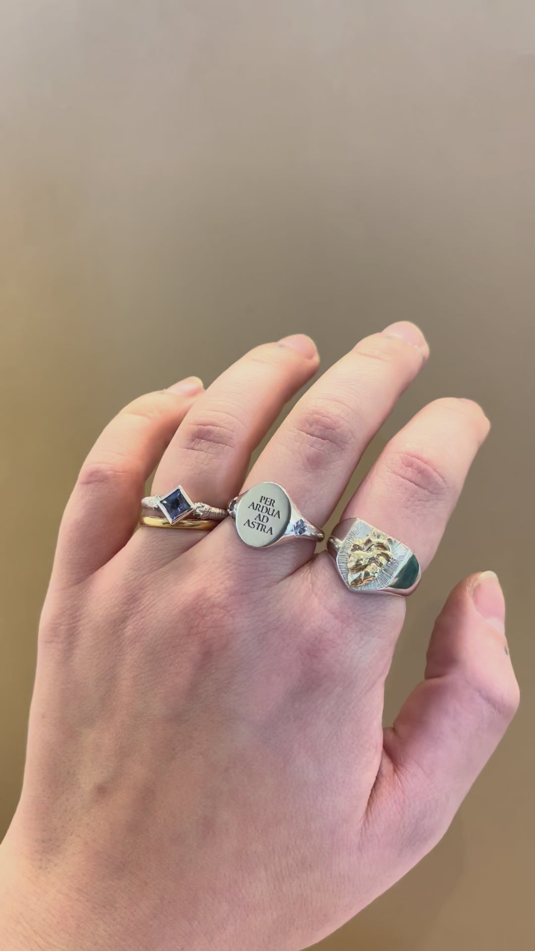 Hand wearing three rings, a silver shield signet with an anatomical human heart in gold, a silver latin motto engraved oval signet ring with birthstone set band, a princess cut gemstone solitaire engagement ring with a blue sapphire with snake head band - emily proudfoot