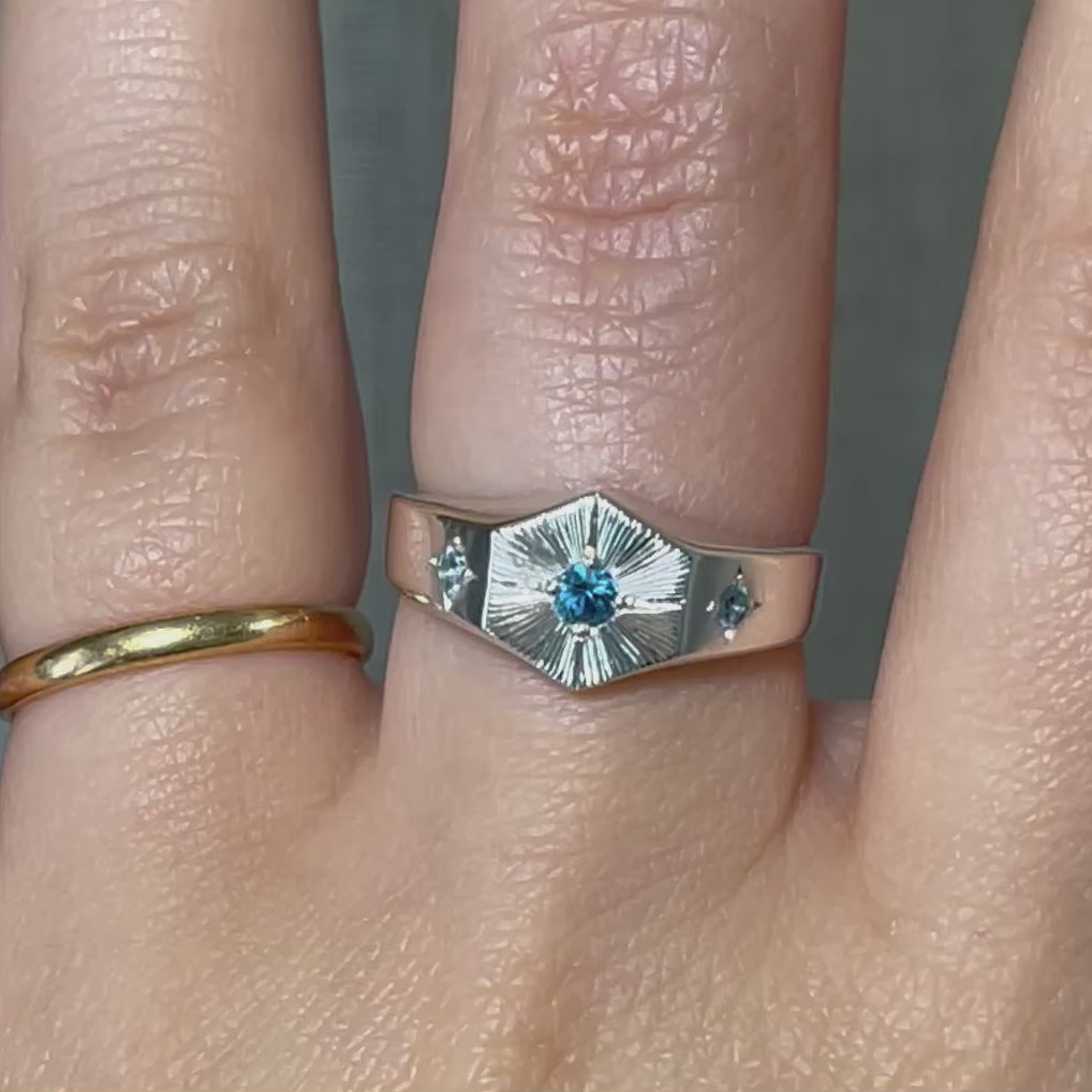 Hexagon-shaped Light Keeper Ring by Emily Proudfoot Jewellery, British jeweller, featuring a 3mm central  blue birthstone in a hand-engraved bright-cut halo, 2mm star-set side stones, polished metal band, with optional interior engraving, symbolising light and guidance.