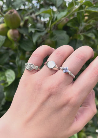 Hand wearing three rings, a silver round signet, a silver hand shake fede ring emerald birthstone set band, a princess cut gemstone solitaire engagement ring with a blue sapphire with snake head band - emily proudfoot