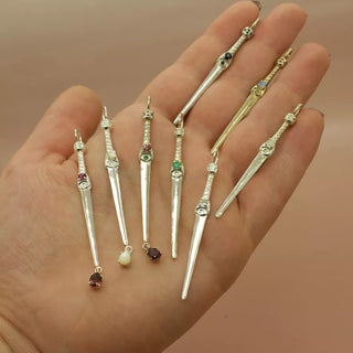 Dagger pendant necklaces in silver and gold, shown on a hand with a pink background